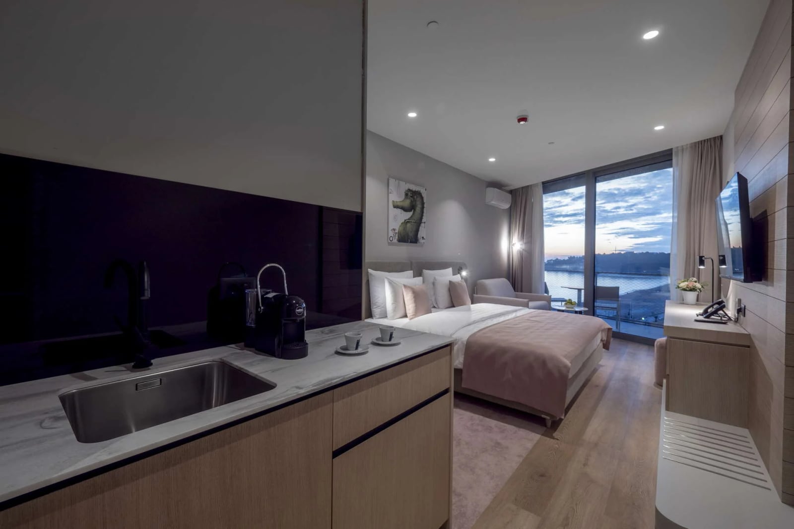 Modern bedroom featuring a bed, kitchenette with sink, and large window overlooking water.
