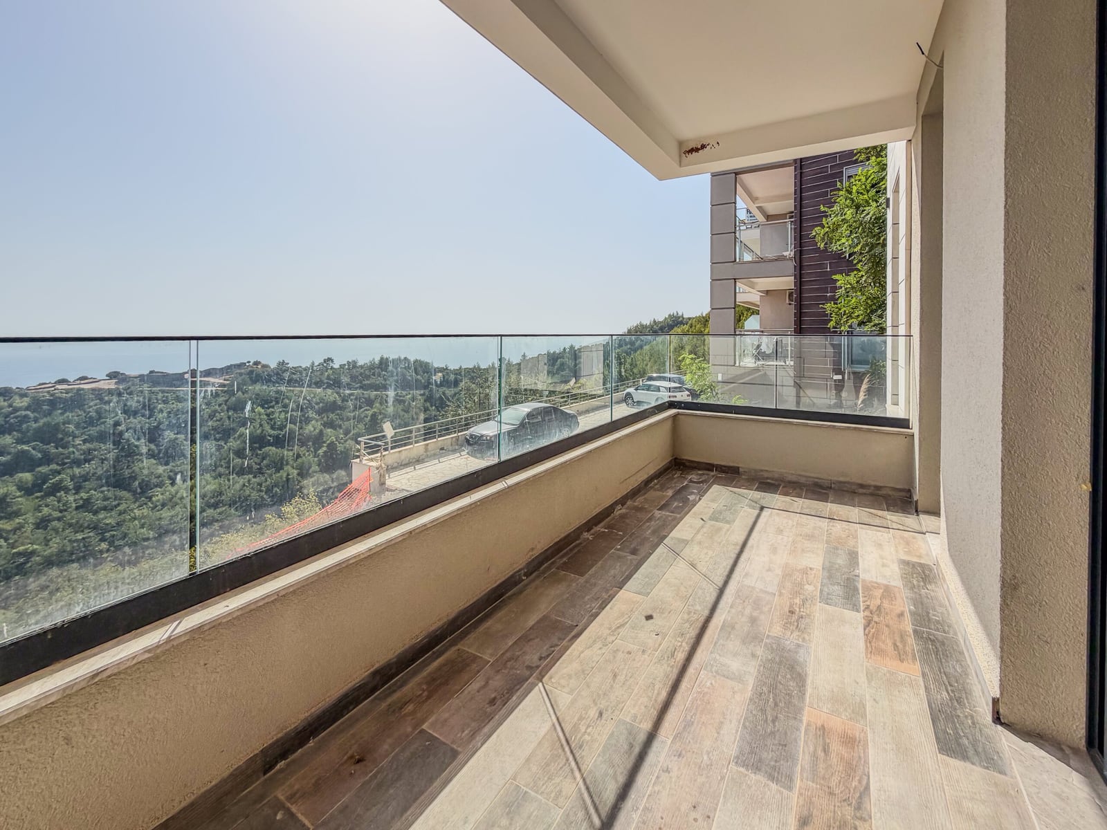 Balcony with wood-effect tiles and glass railing overlooking a forested hillside and sea.