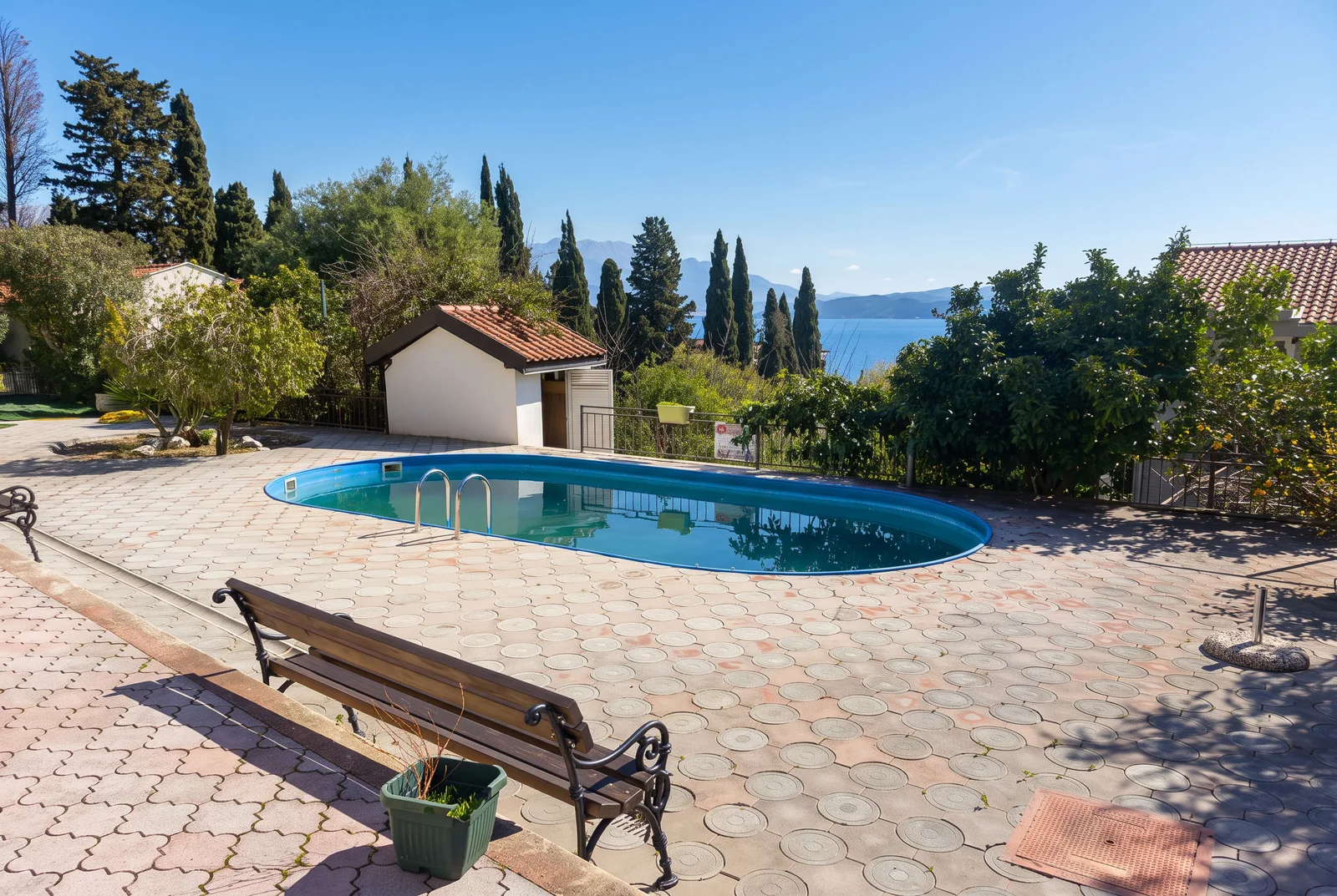 Outdoor pool on a paved terrace with a wooden bench, surrounded by trees and a sea view.