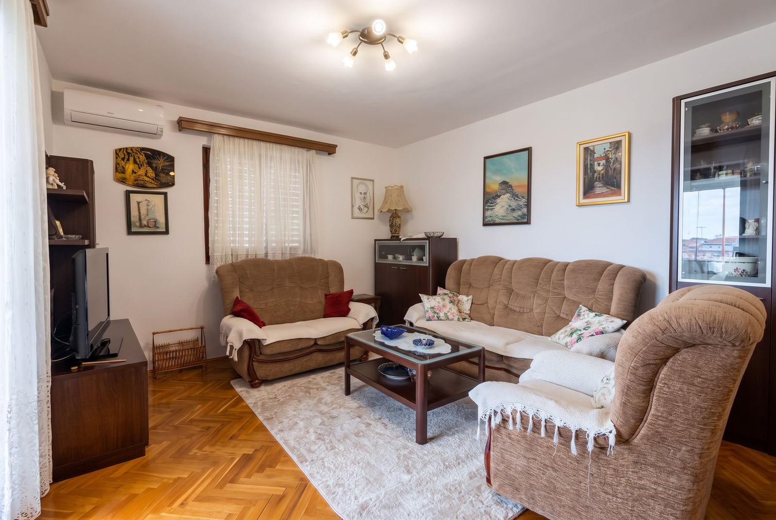 Living room featuring brown sofas, parquet flooring, a TV unit, coffee table, and framed artwork on white walls.