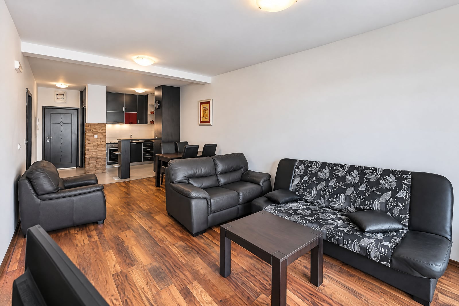 Living room with black leather sofas, wooden flooring, and an open kitchen area visible in the background.