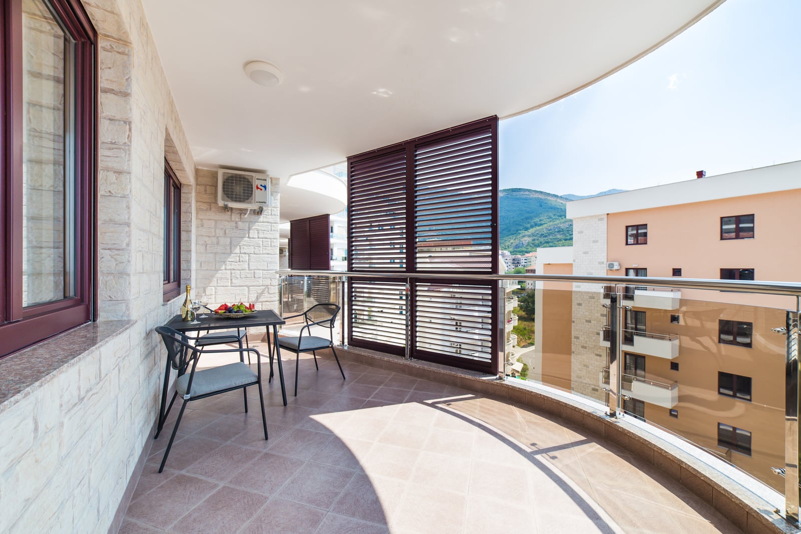 Spacious balcony with furniture and mountain views in a Becici apartment, Montenegro: stone finishes and glass railings.
