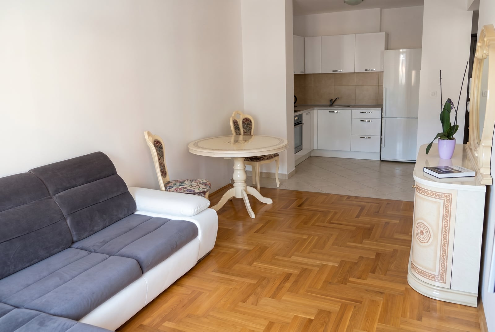Living room with parquet flooring, a sofa, and a dining area in an apartment in Budva, Montenegro. White kitchen with appliances in the background.