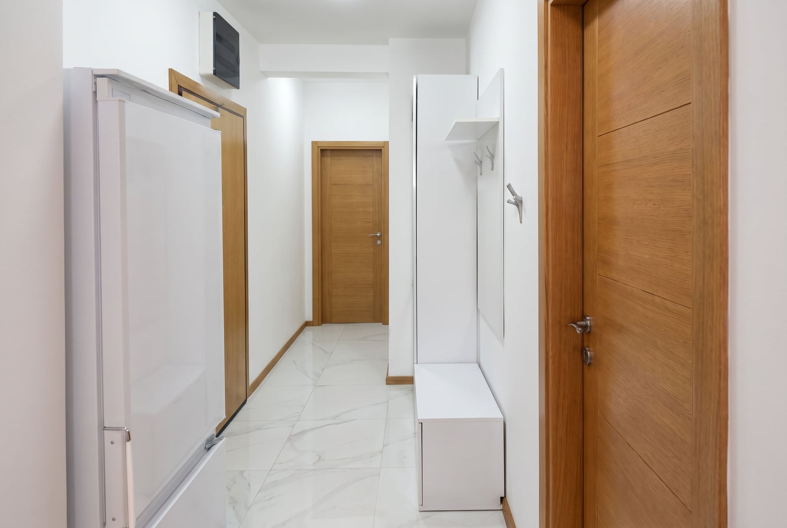 Narrow hallway with white walls, marble floor, wooden doors, and a white entryway bench with coat hooks.