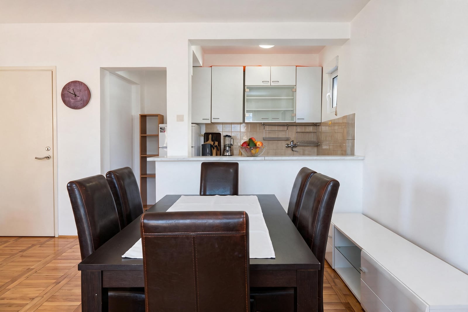 Dining table with brown leather chairs in front of a kitchen area featuring white cabinets and tiled backsplash.