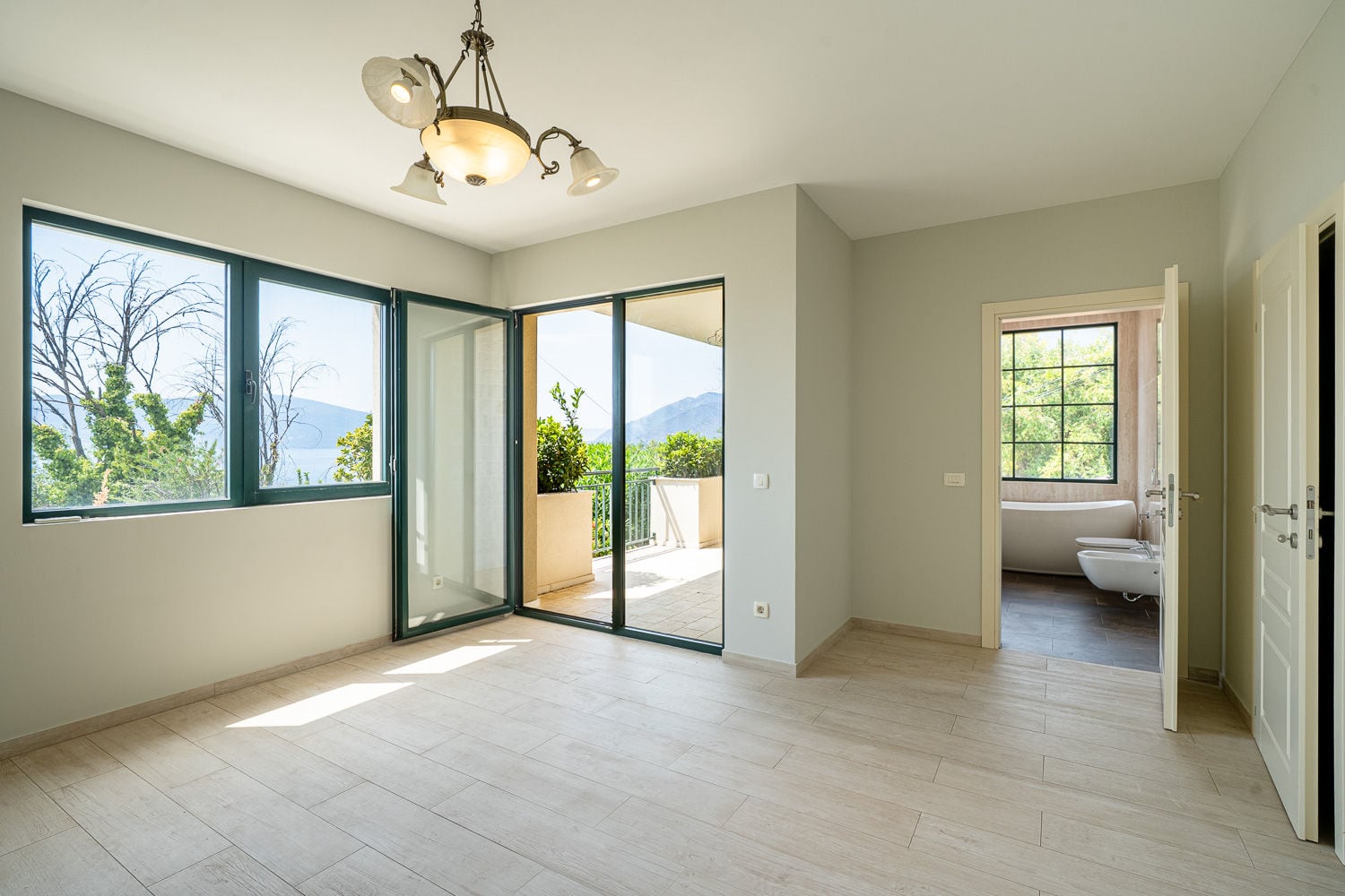 Empty room with wood-look tile floor, balcony access, water view, and an adjacent bathroom visible through a doorway.