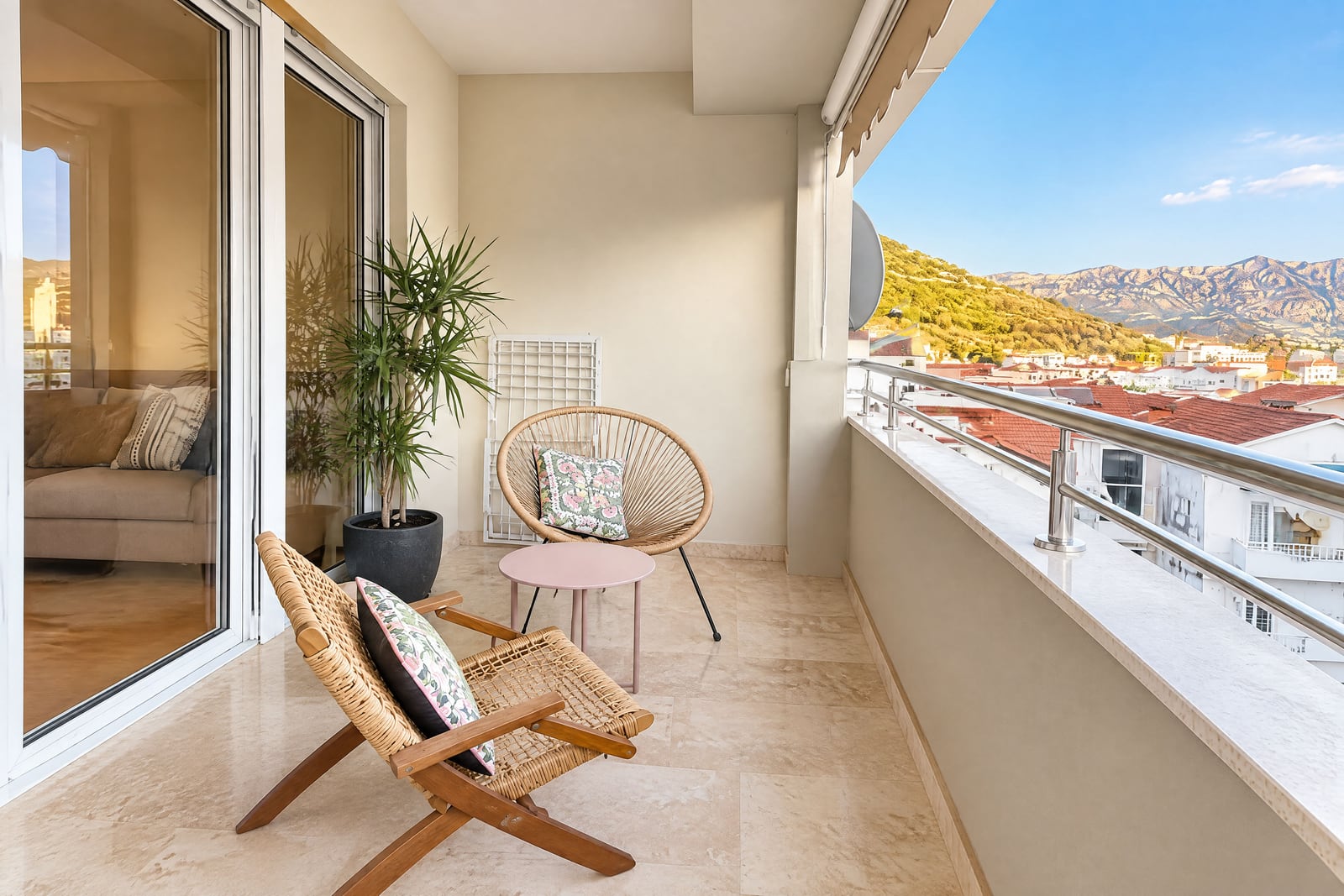 Balcony with wicker chairs, small table, potted plant, and a view over rooftops to mountains.