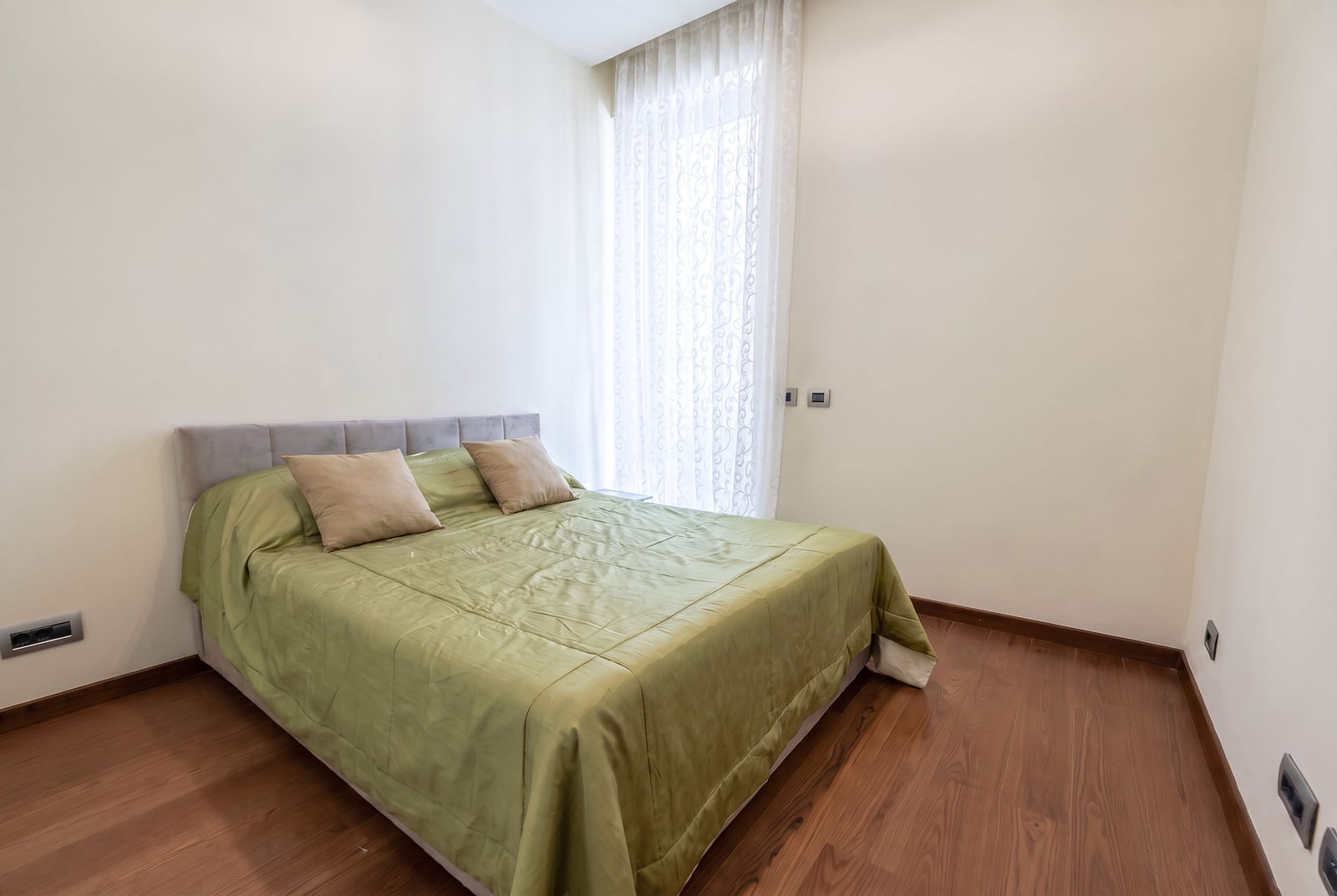 Bedroom featuring a double bed with green bedding, wood flooring, and sheer white curtains by the window.