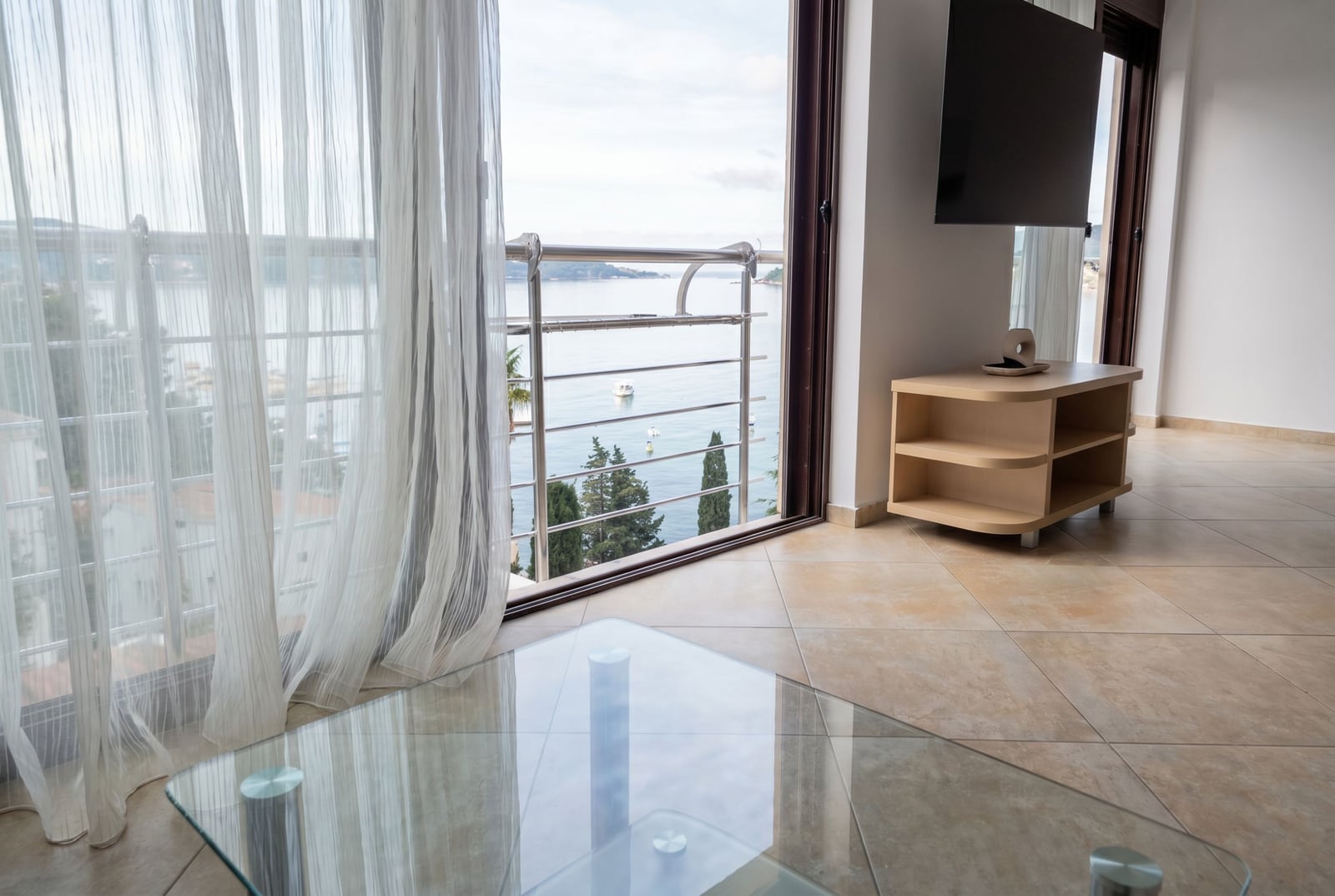 Living room with panoramic sea view in an apartment, Rafailovici, Montenegro: glass table, TV, and access to the balcony.