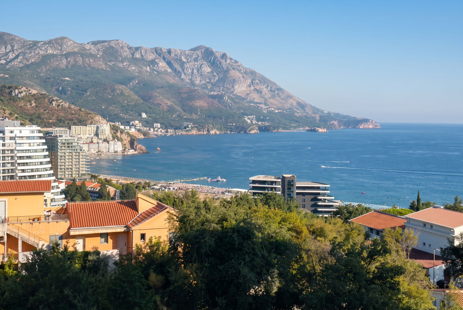 Panoramic view of the sea coast, mountains, and city buildings with red roofs amidst greenery.