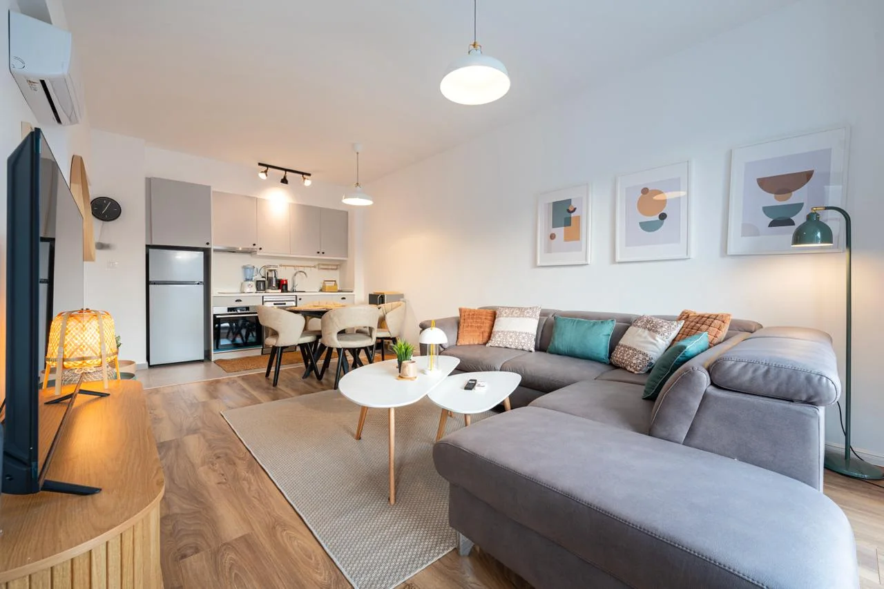Modern living room with a grey sectional sofa, white tables, and an open kitchen in the background.