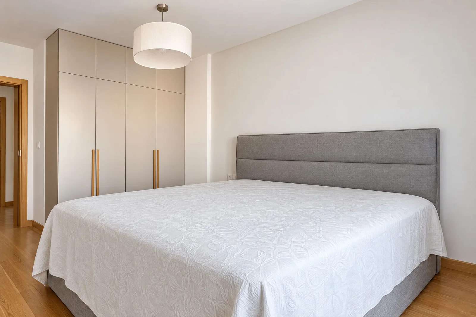 Modern bedroom featuring a large bed with a grey upholstered headboard, white patterned coverlet, and built-in wardrobe with gold handles.