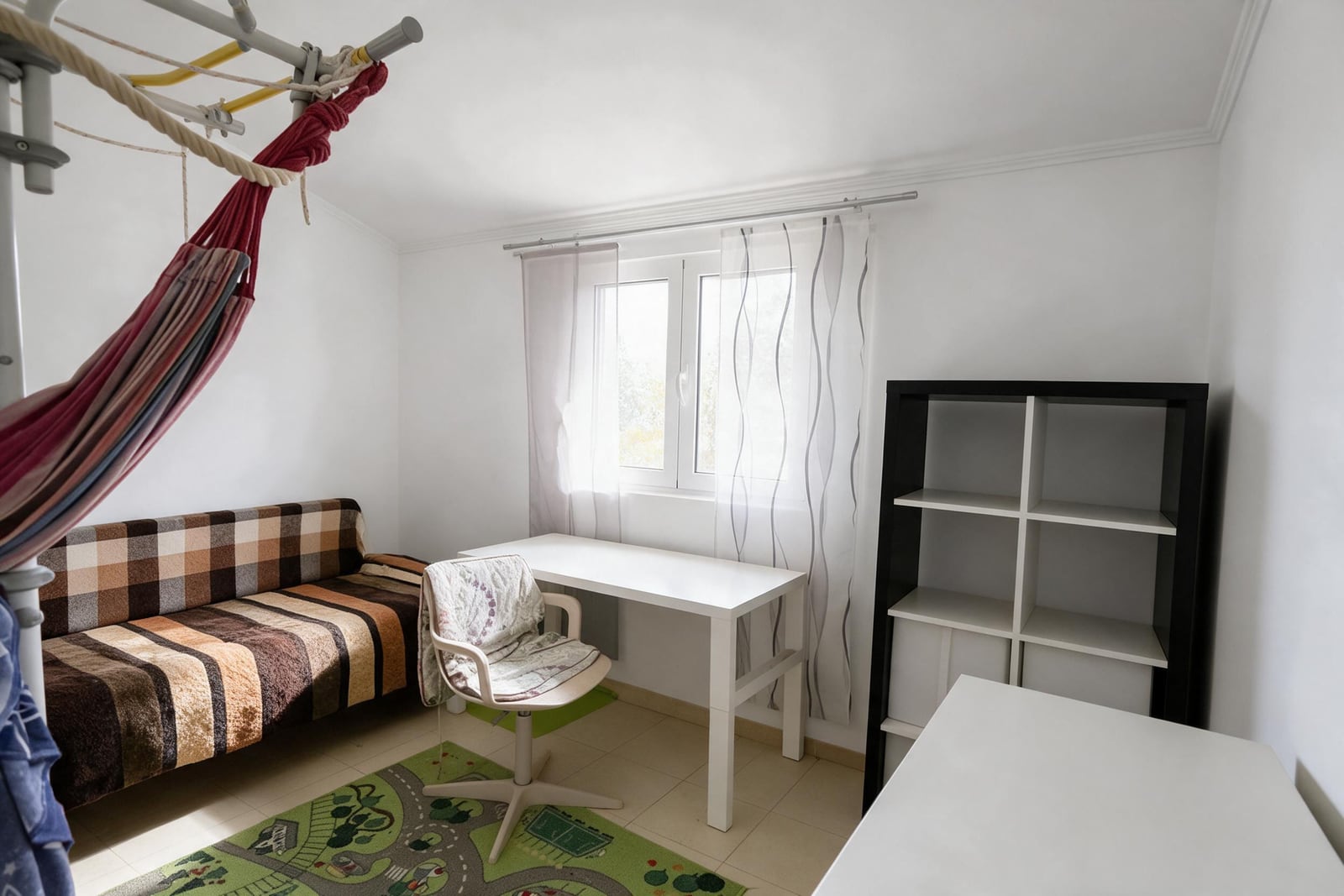 Room with plaid sofa, white desk, black shelf, hammock, and children's play rug.