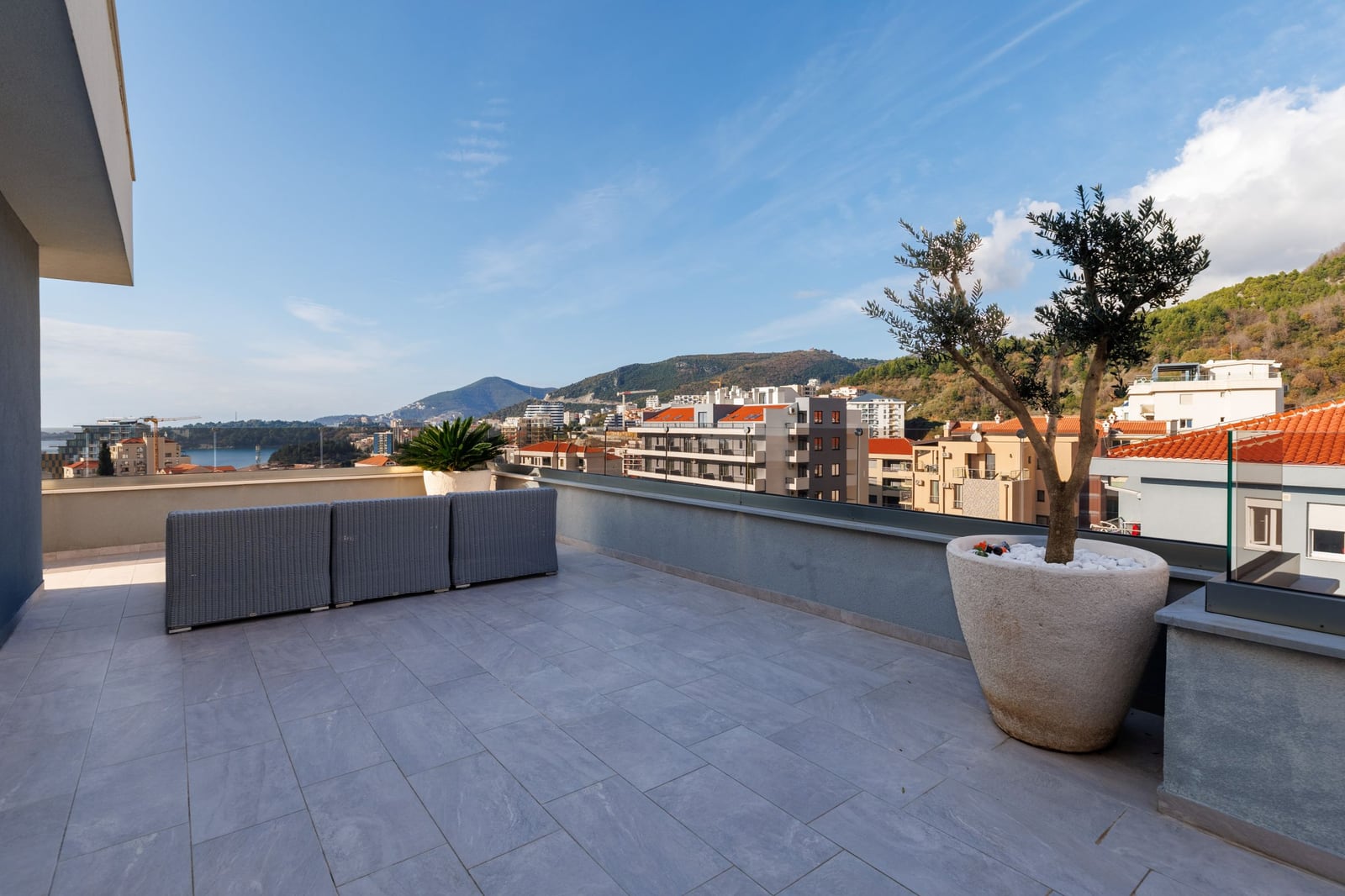 Spacious penthouse terrace in Becici, Montenegro, featuring wicker furniture, an olive tree, and panoramic sea views.