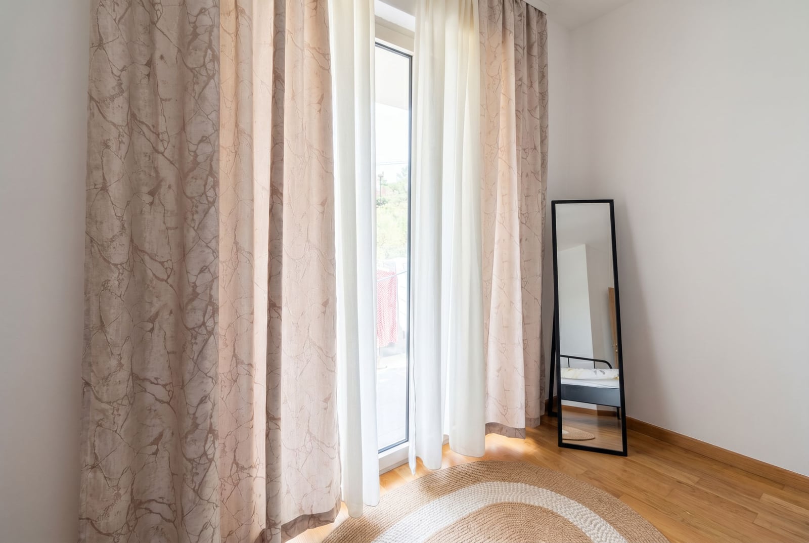 Bedroom in an apartment in Bečići, Montenegro: panoramic window with beige curtains, a floor mirror, and a woven rug.