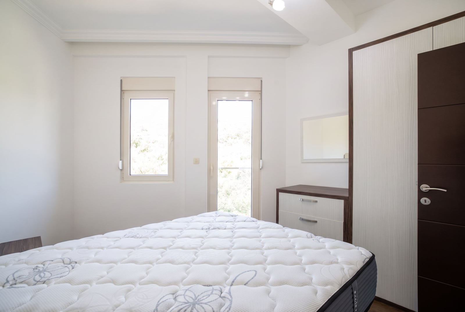Bright bedroom featuring a white mattress, wooden wardrobe, and dresser near the window.