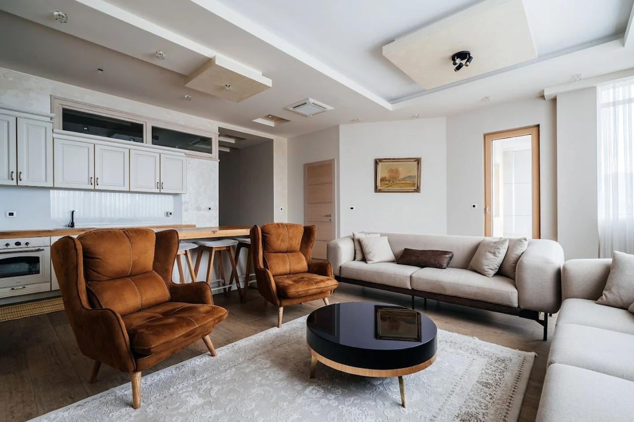 Modern living room with kitchen in an apartment in Budva, Montenegro: beige sofas, brown armchairs, and a round table.