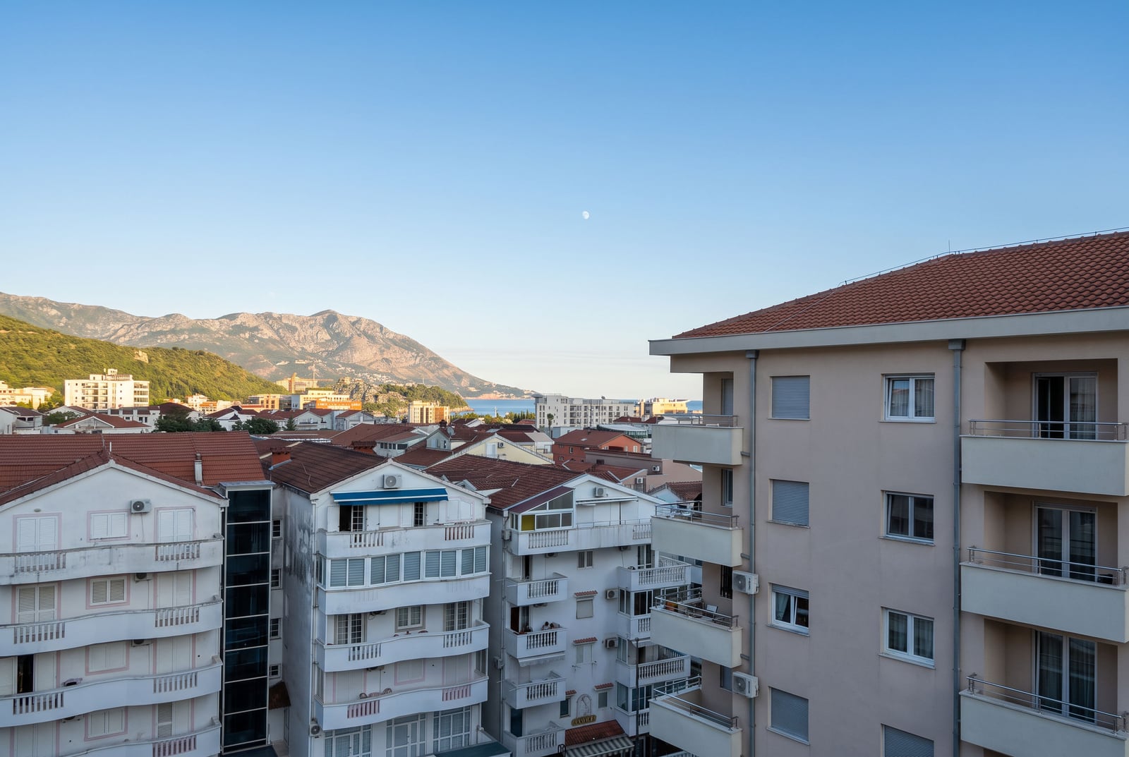 View of city rooftops, multi-story buildings, and mountains under a clear blue sky with a visible moon.