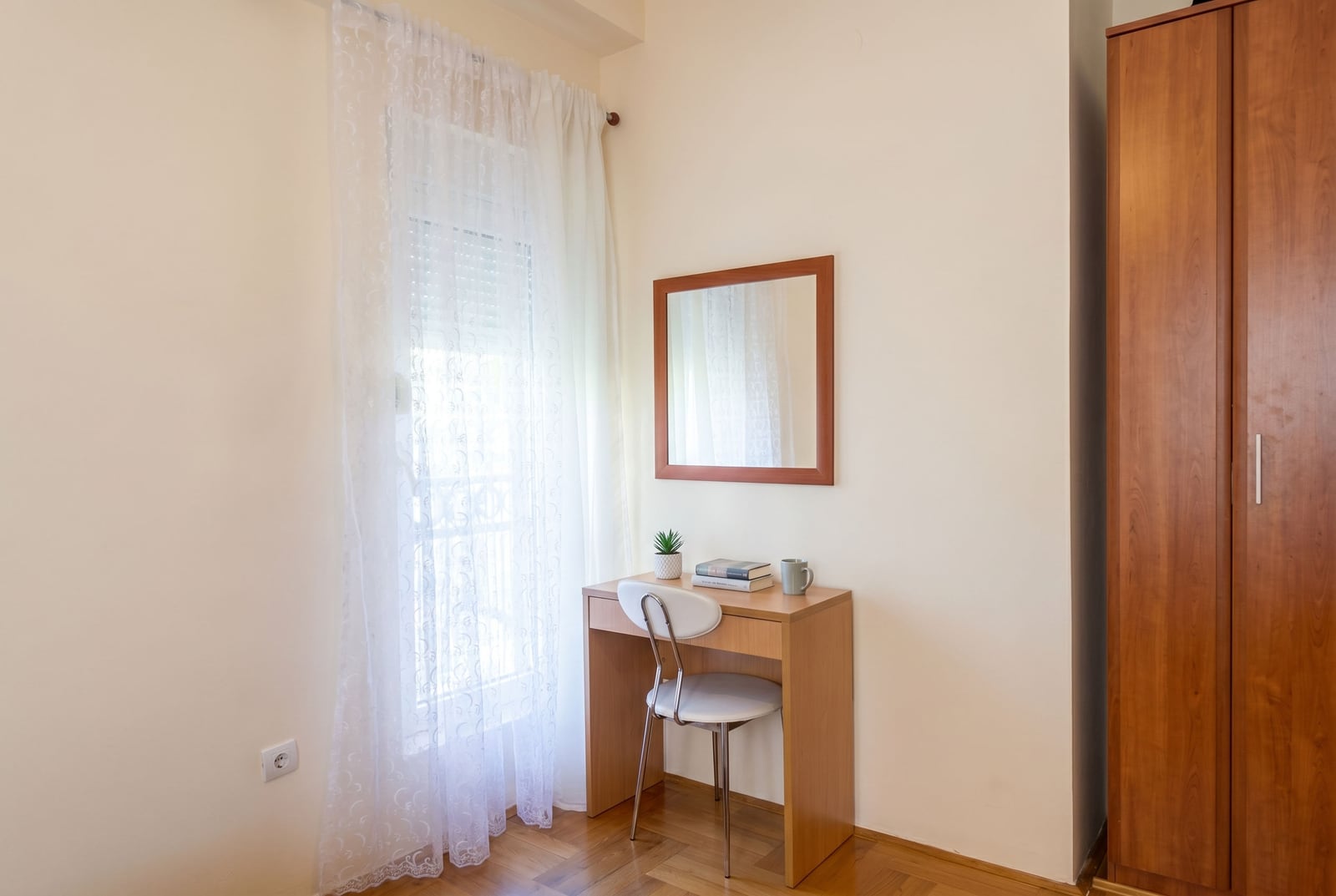 Cozy bedroom with a desk and wardrobe in a Budva apartment, Montenegro: bright interior, parquet flooring, and a window with sheer curtains.