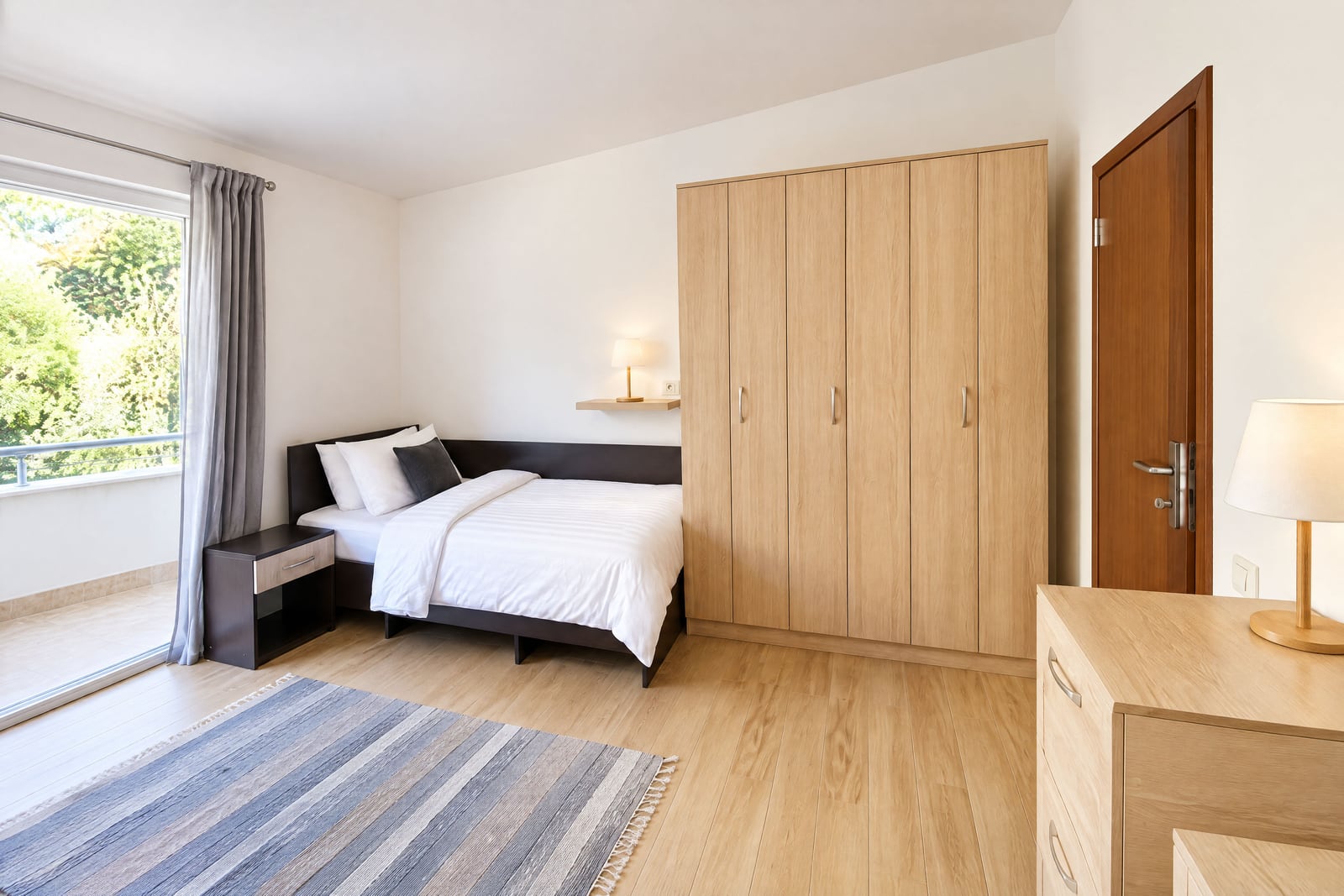 Bright bedroom featuring a single bed, large light wood wardrobe, striped rug, and sliding glass door.