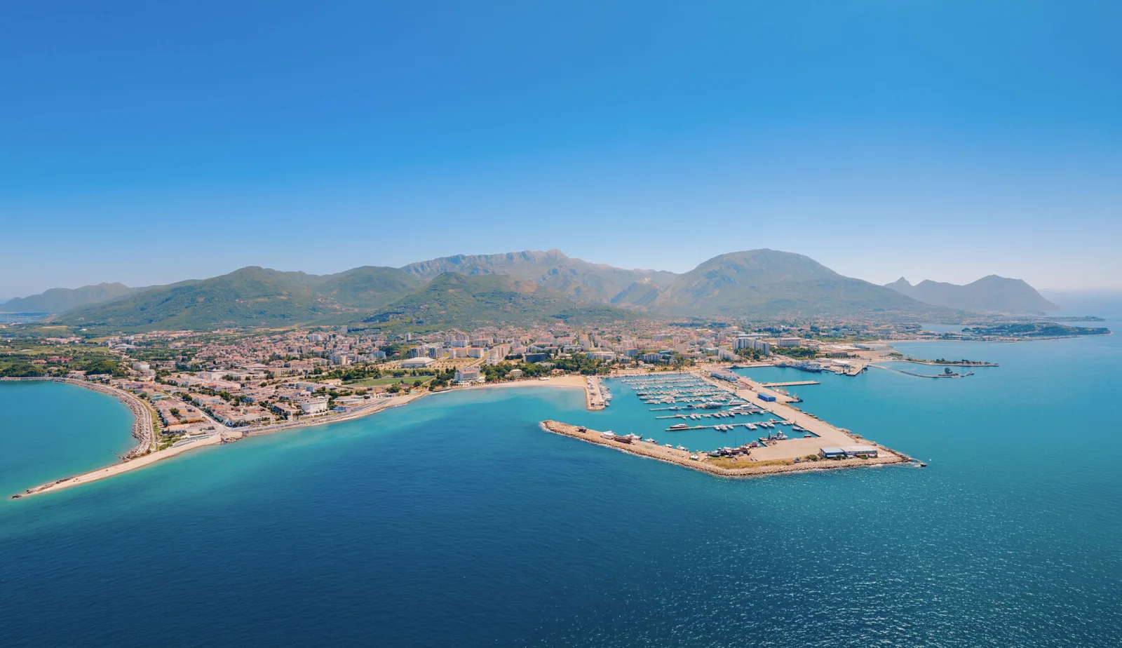 Aerial view of a coastal city featuring a marina, beach, and mountains in the background.