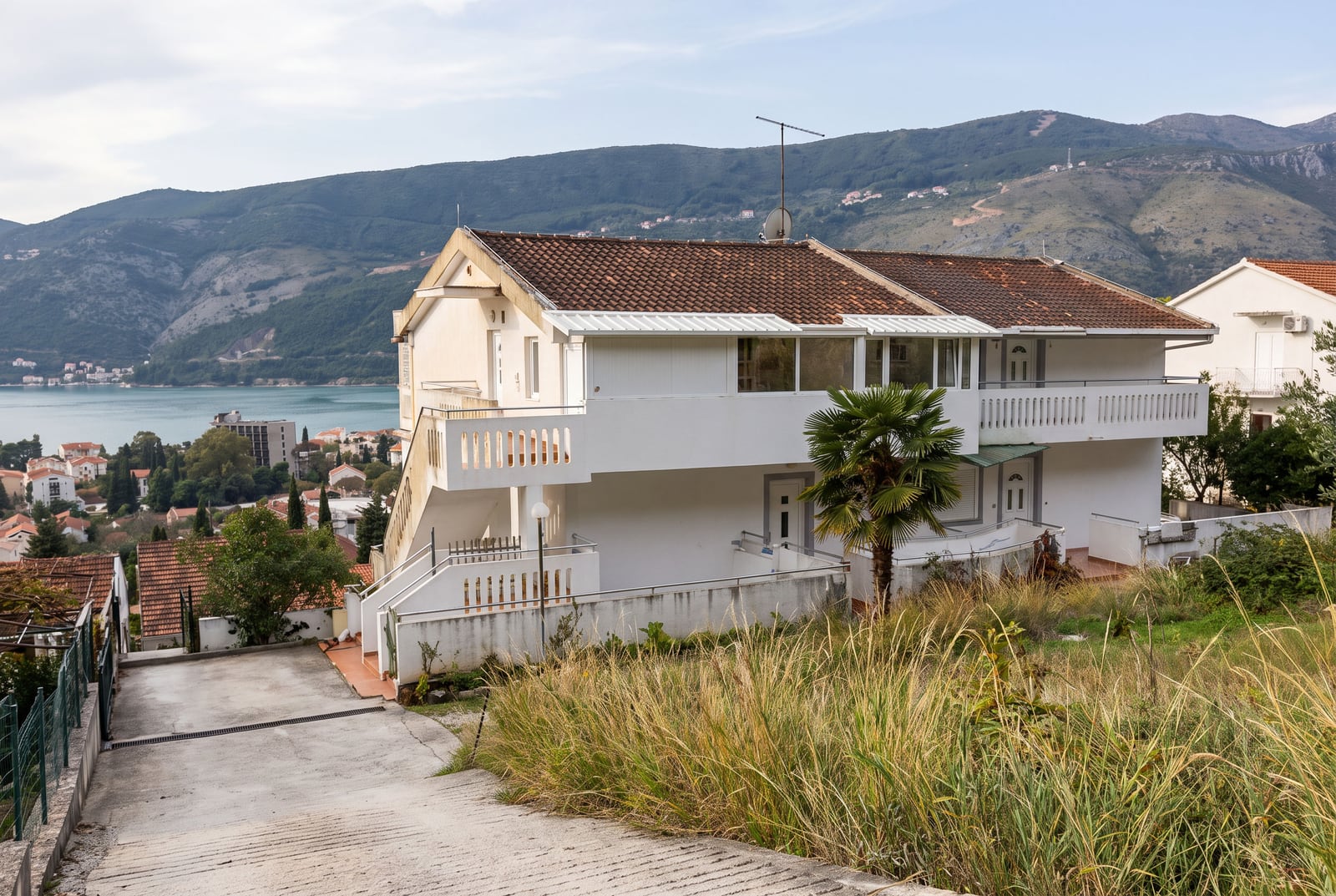 Exterior view of a white two-story house with a tiled roof and balcony, situated on a hillside near the sea with mountains in the background.