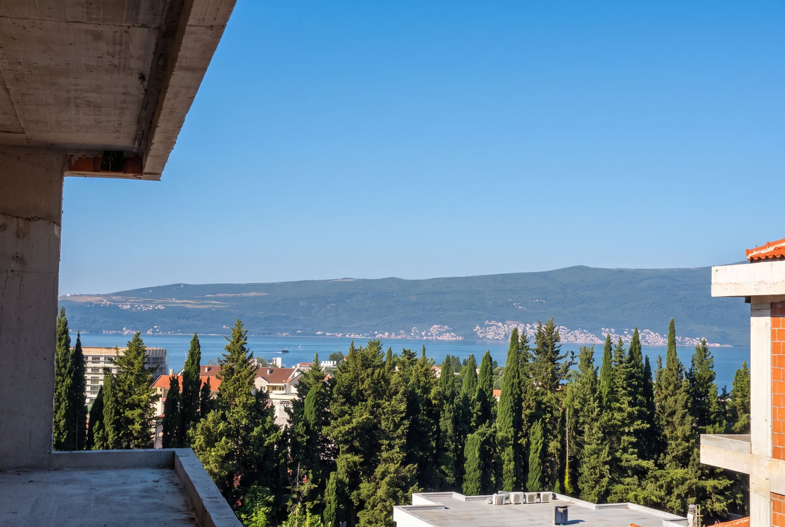 Concrete balcony overlooking sea, cypress trees, and mountains under clear sky.
