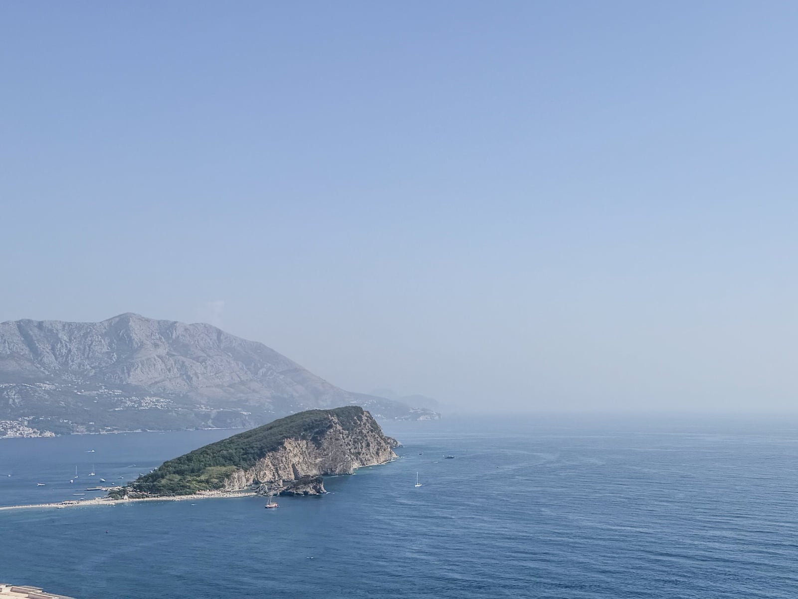 Panoramic view of the sea, a rocky green peninsula, and mountains in the background under a clear sky.