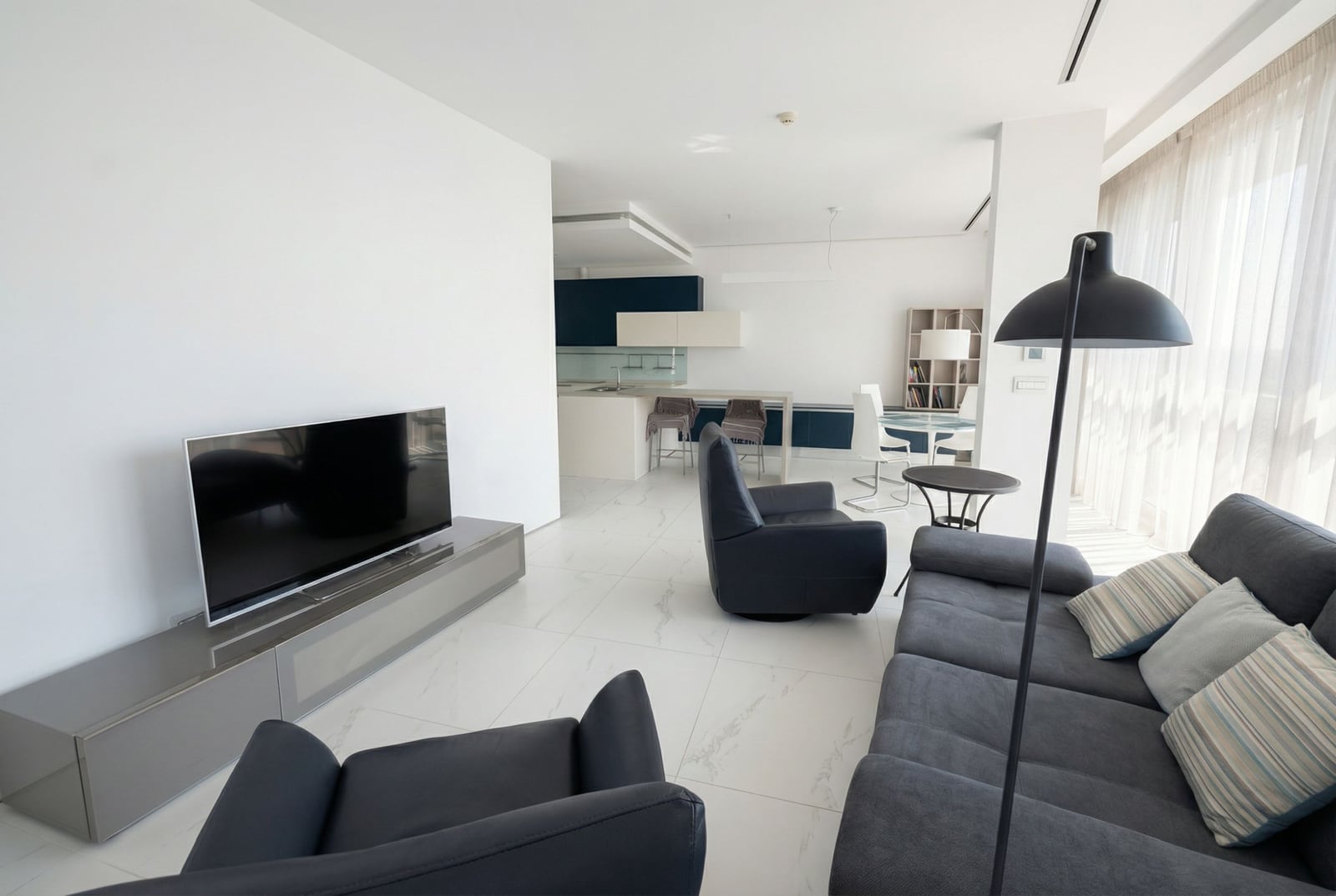 Modern living room with kitchen in Dukley apartments, Budva, Montenegro: gray sofa, armchairs, and panoramic windows.