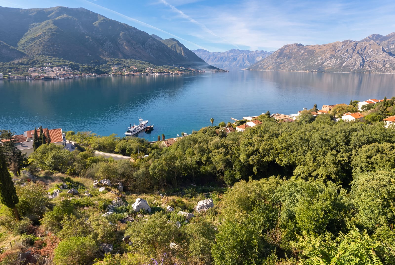 Panoramic view of a bay, mountains, and a village with red roofs amidst greenery.