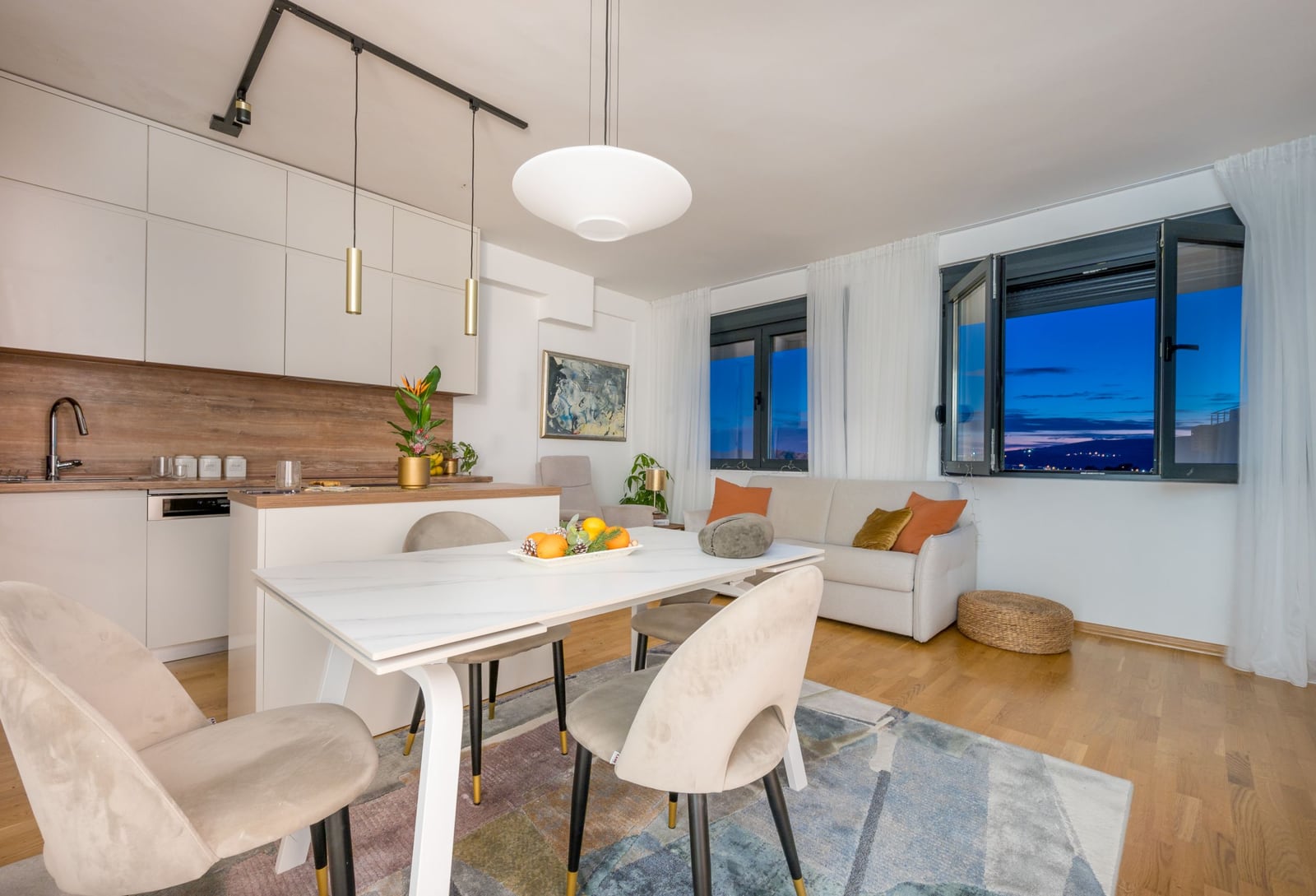 Modern living room with a kitchen in an apartment in Tivat, Montenegro: light-colored furniture, dining area, and a view of the city at night.