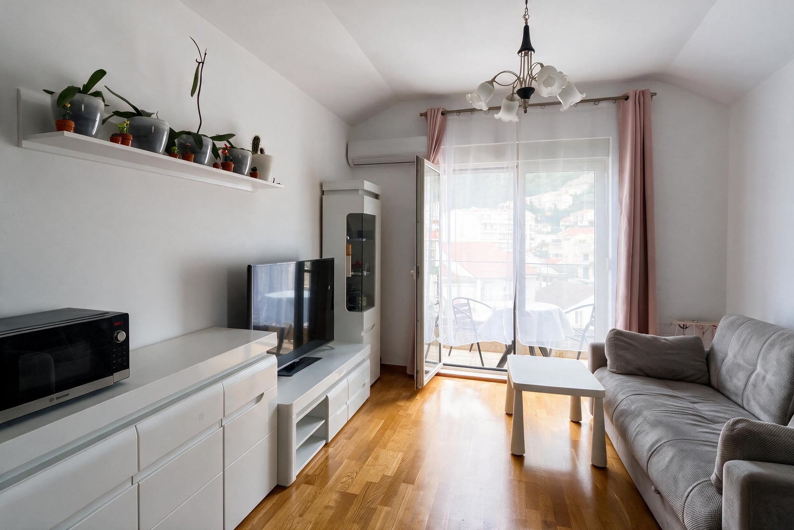 Living room with grey sofa, white TV unit, microwave, wooden floor, and glass door leading to a balcony.