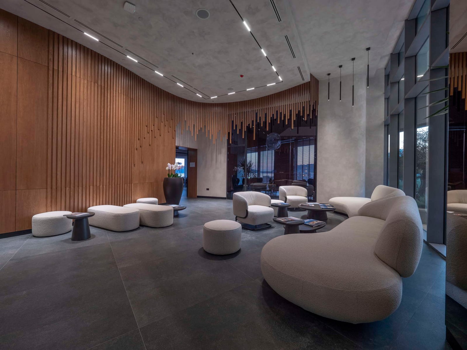 Modern lobby with curved wood slat wall, beige curved sofas, and dark tiled flooring under linear ceiling lights.