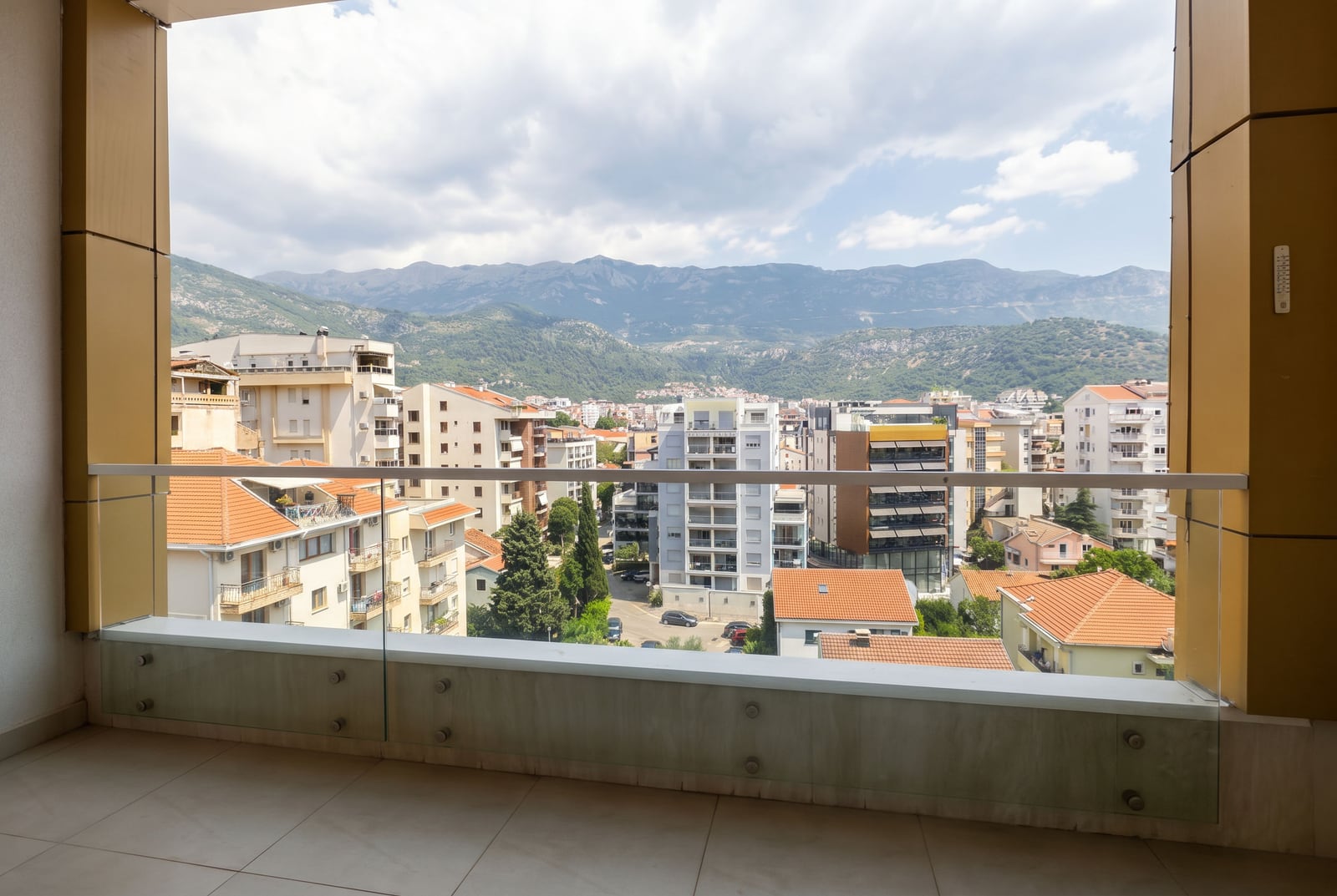 View from the balcony of a modern apartment in Budva, Montenegro: a panorama of the city, residential buildings, and a mountain landscape in the background.