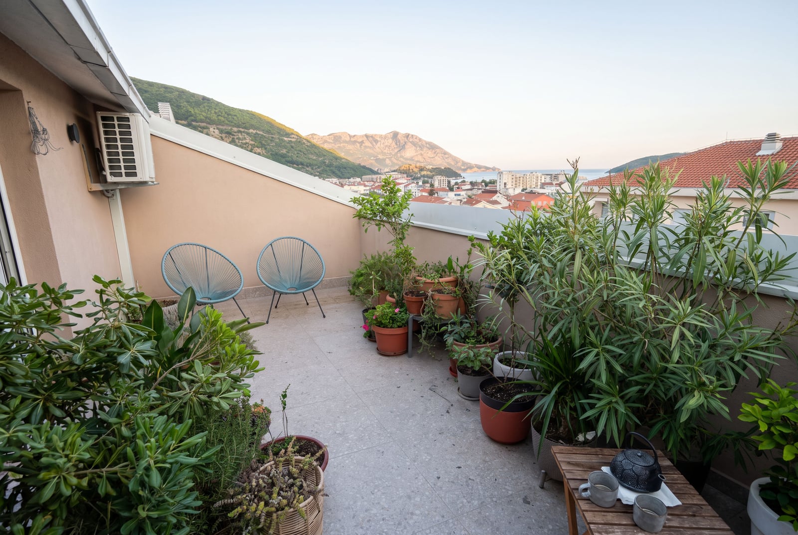 Balcony with blue chairs, many potted plants, and a view of mountains and the sea.