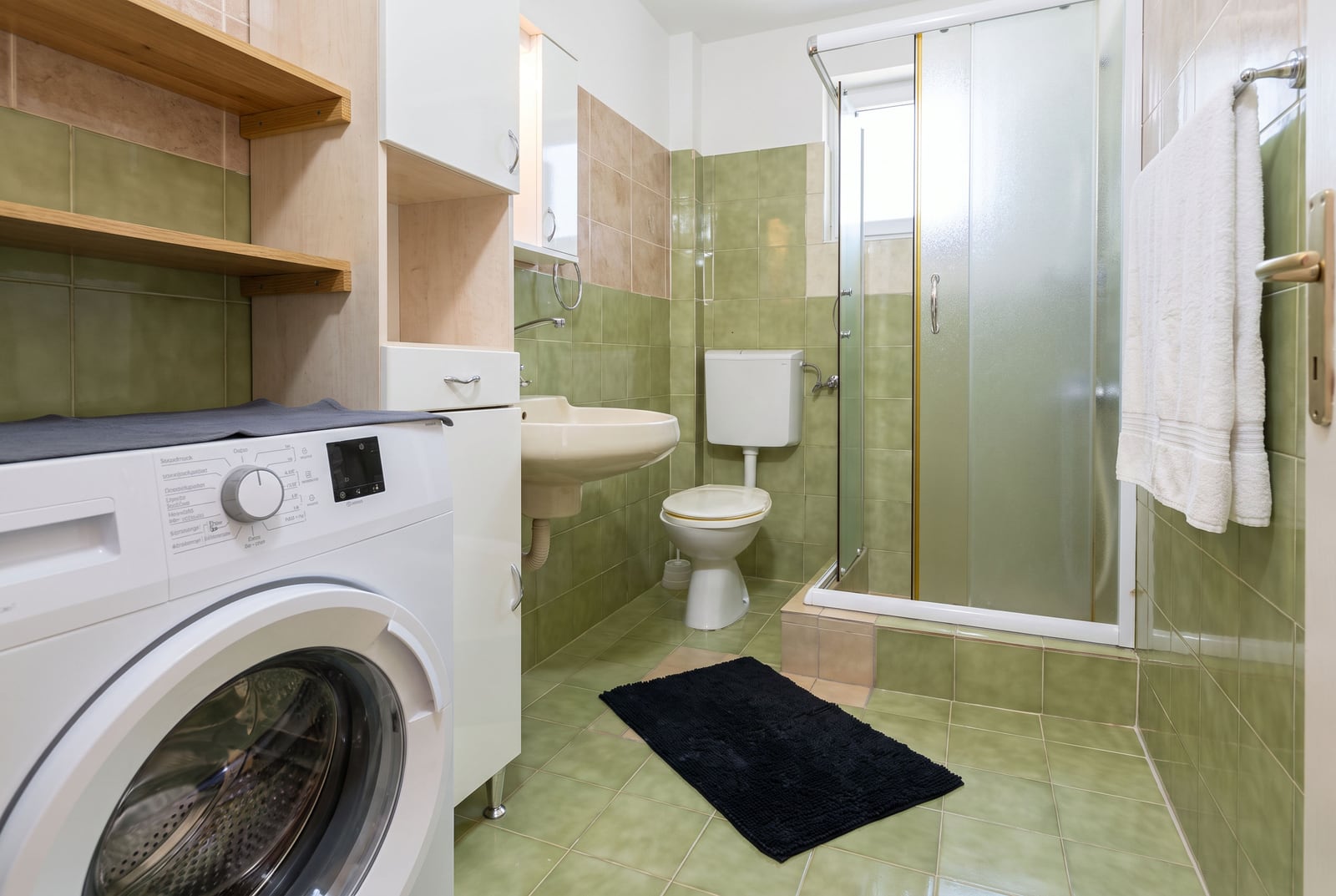 Bathroom with green tiles, washing machine, toilet, sink, and glass shower enclosure visible.
