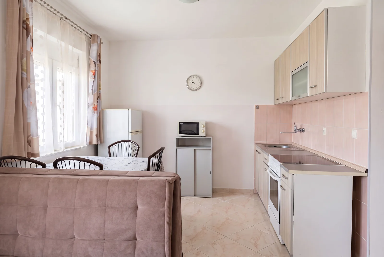 Kitchen with light cabinets, pink tiles, fridge, microwave, dining table, and sofa back visible in foreground.