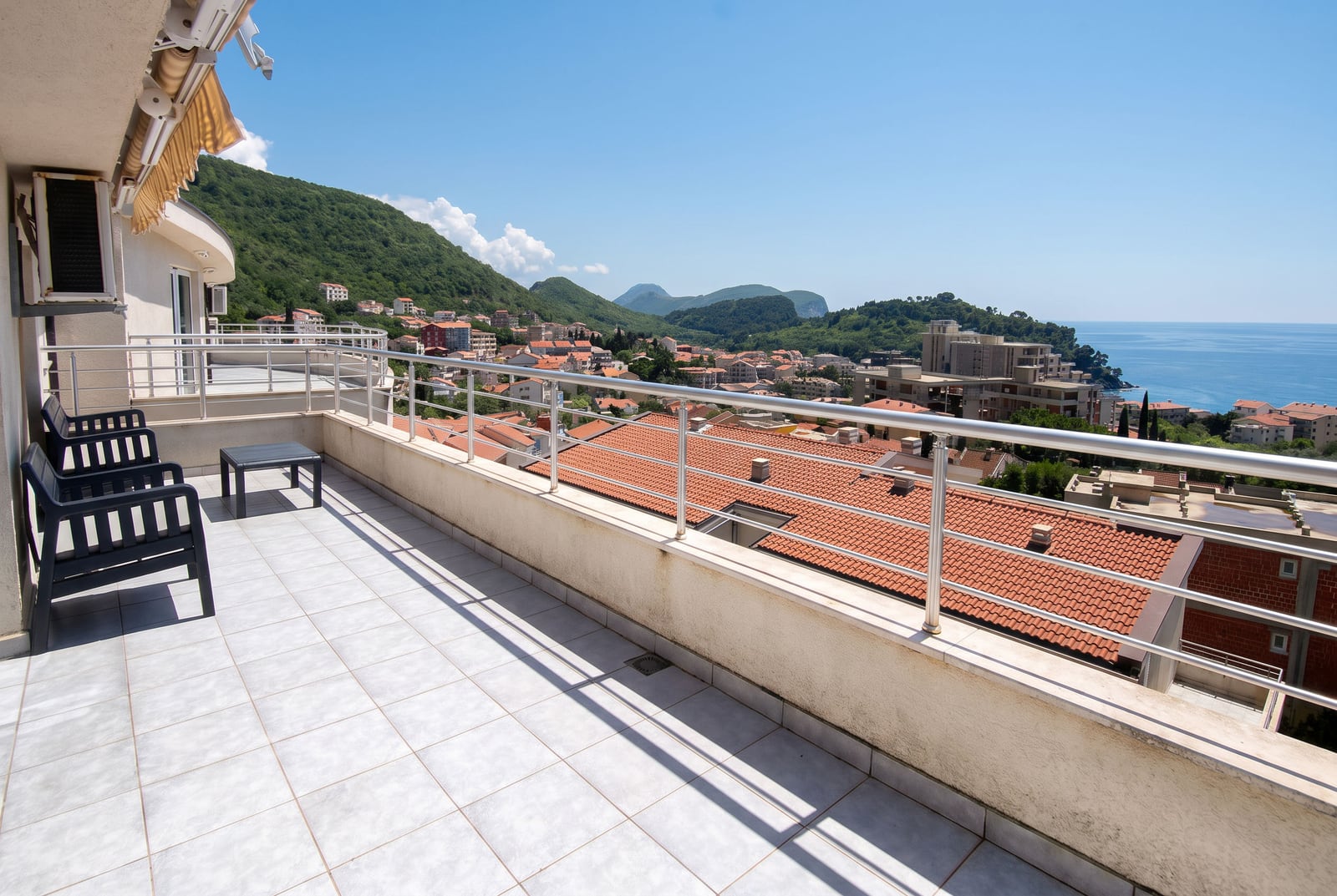 Wide tiled balcony with black plastic chairs overlooking a coastal town, green hills, and the sea.