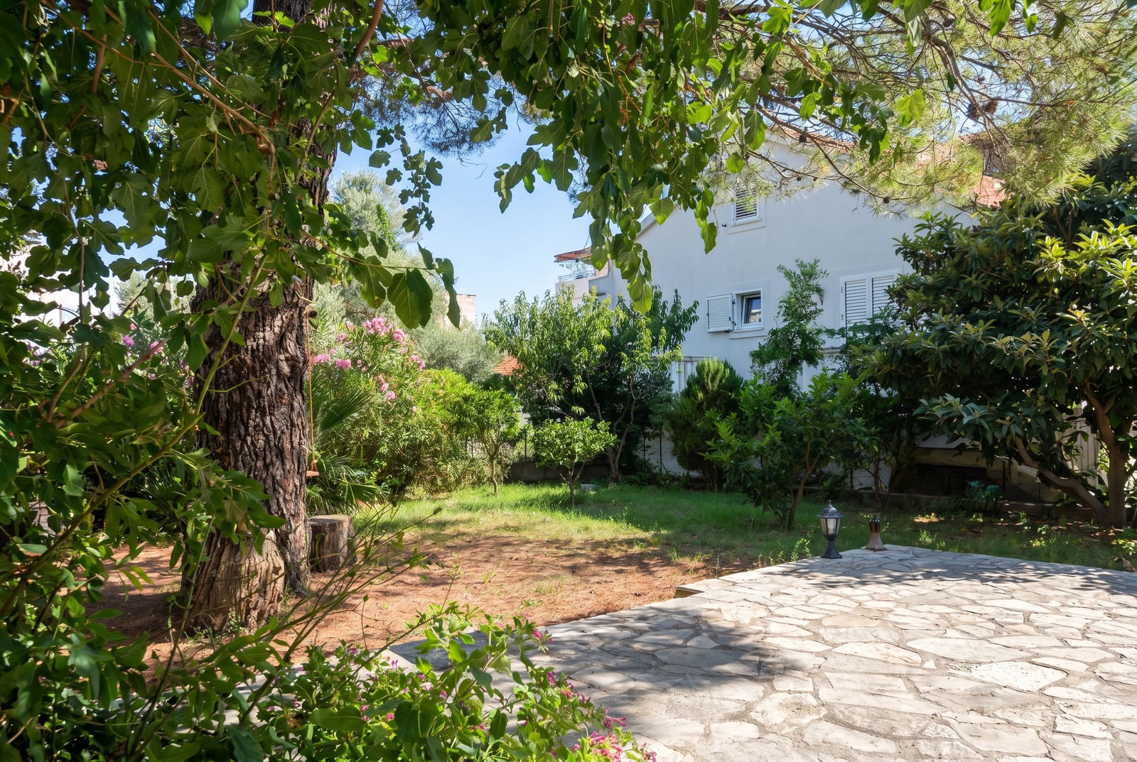 Cozy green courtyard with a stone terrace by a house in Budva, Montenegro, surrounded by trees and flowering shrubs.