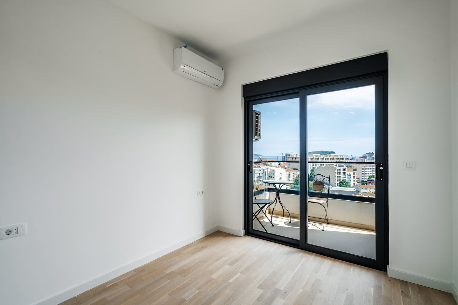 Empty room with light wood flooring and AC unit, featuring sliding doors leading to a balcony with city view.