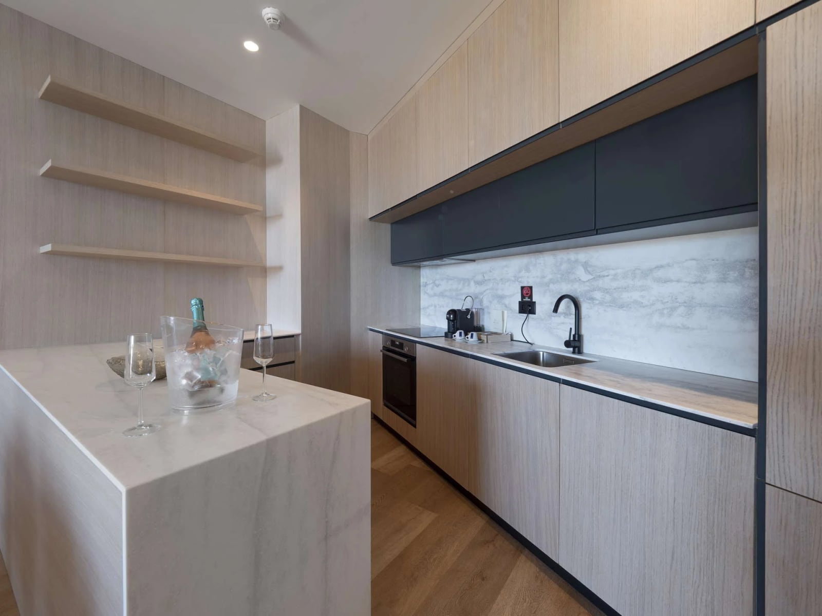 Modern kitchen with light wood cabinets, marble countertops, a sink, built-in oven, and open shelving.