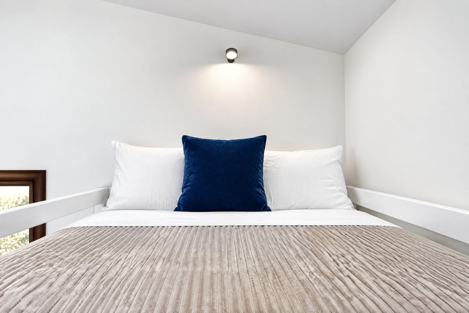 Bed with beige coverlet, white and blue pillows against a white wall under a spot light.