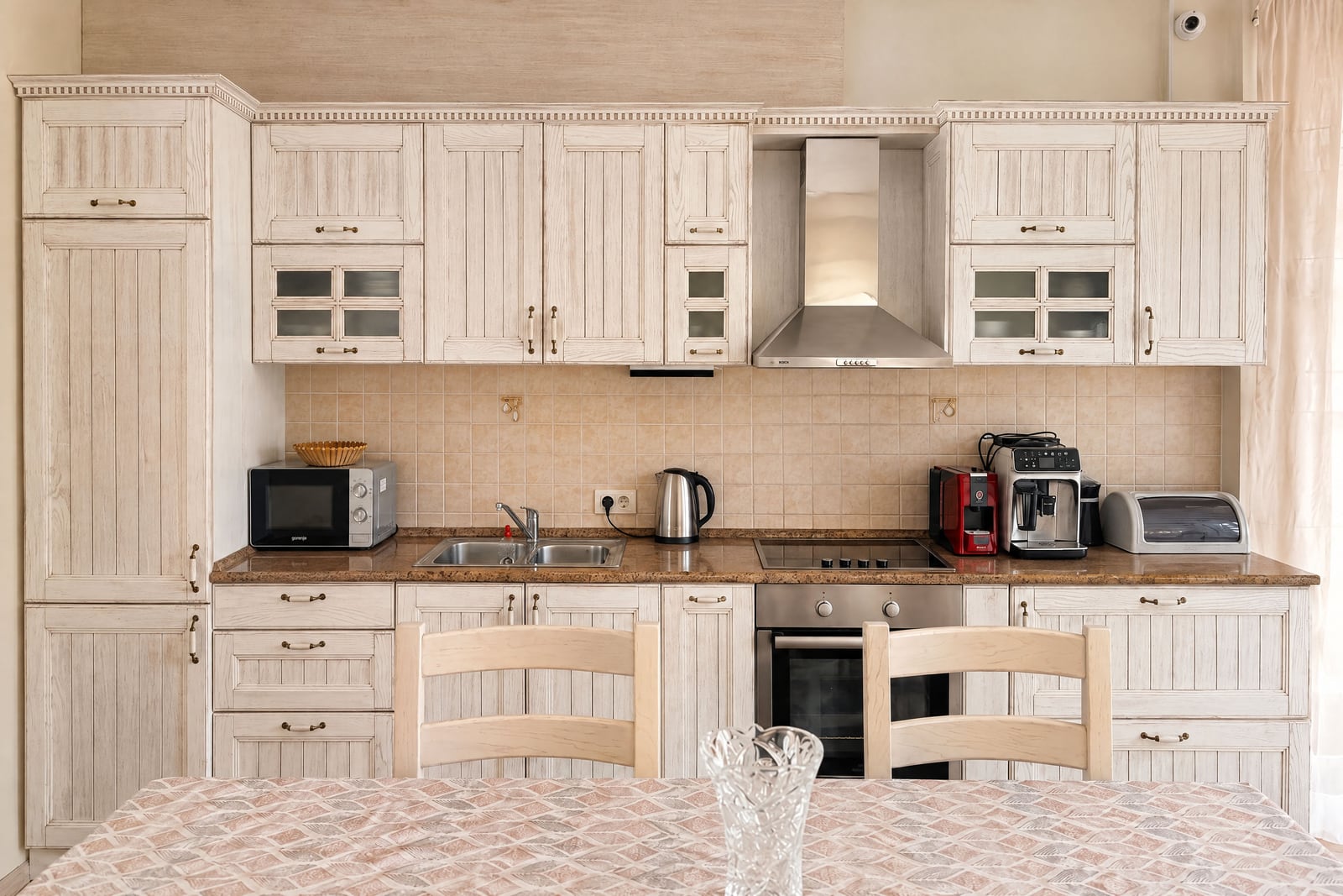 Bright kitchen with white wooden cabinets, stainless steel hood, stove, oven, microwave, and a dining table in the foreground.