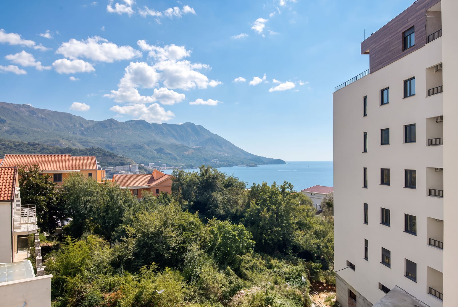 View of the sea and mountains from a high-rise building balcony surrounded by trees and houses.
