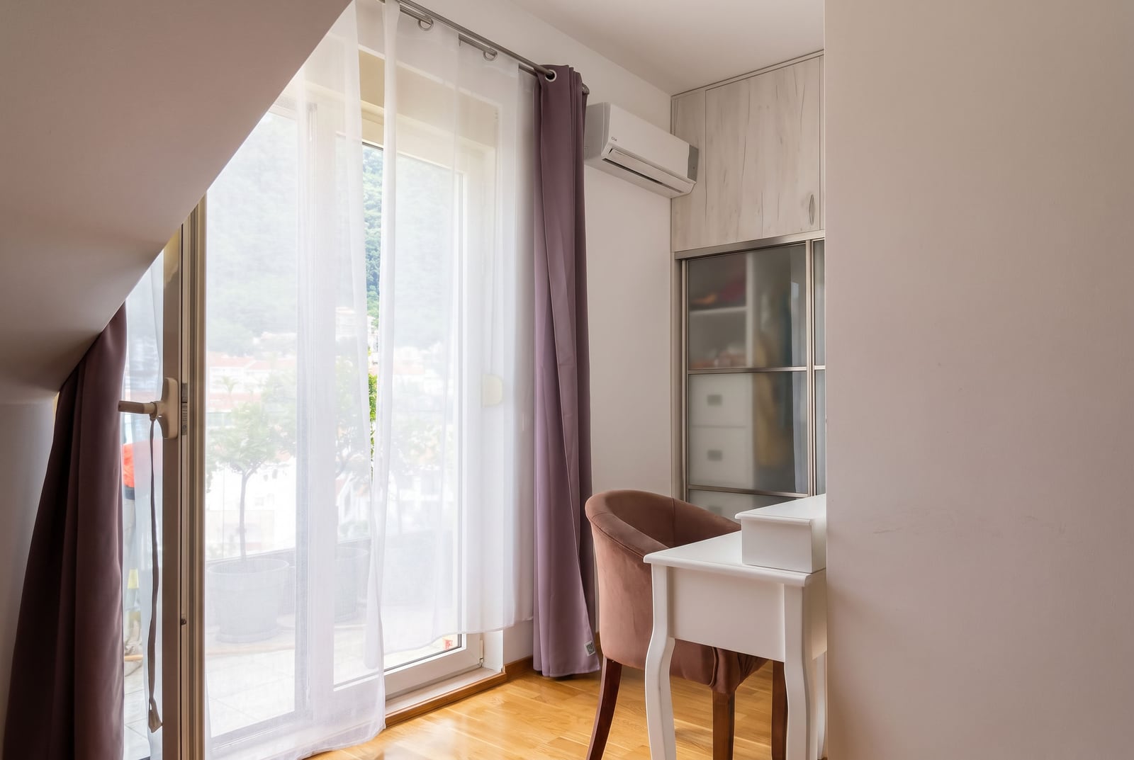 Room with a white desk, pink chair, wardrobe, and balcony featuring purple curtains.