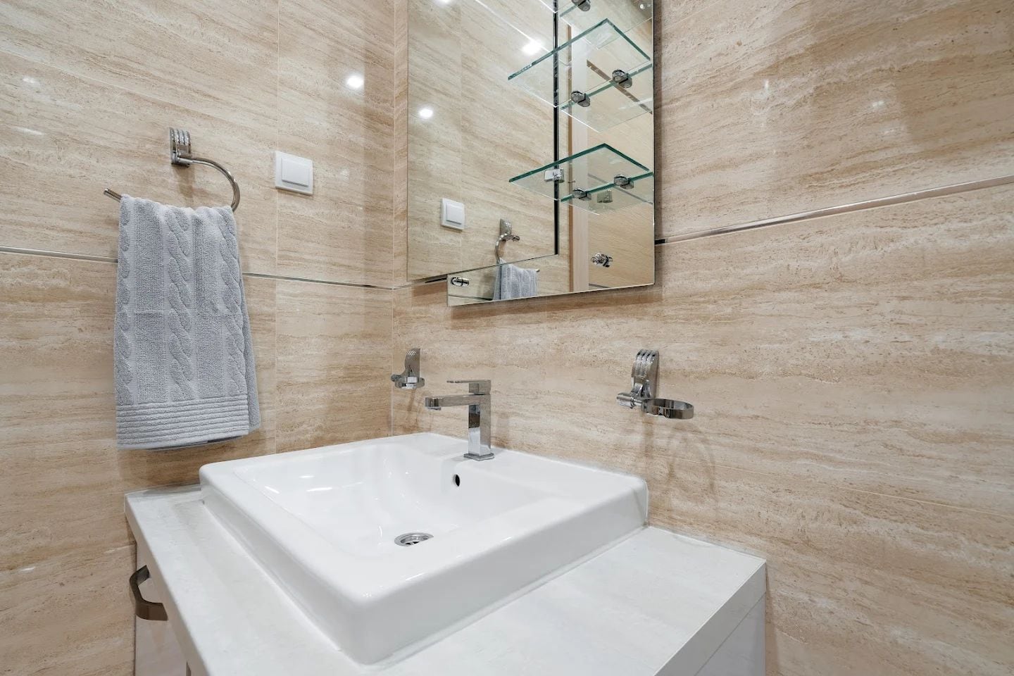Modern bathroom in an apartment in Budva, Montenegro: white sink, mirror with shelves, and beige tile finish.