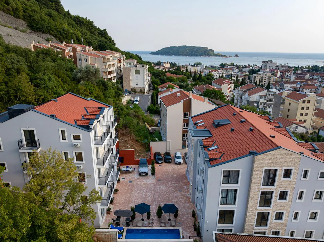 Aerial view of residential buildings with red roofs, a parking area, pool, and sea with an island.