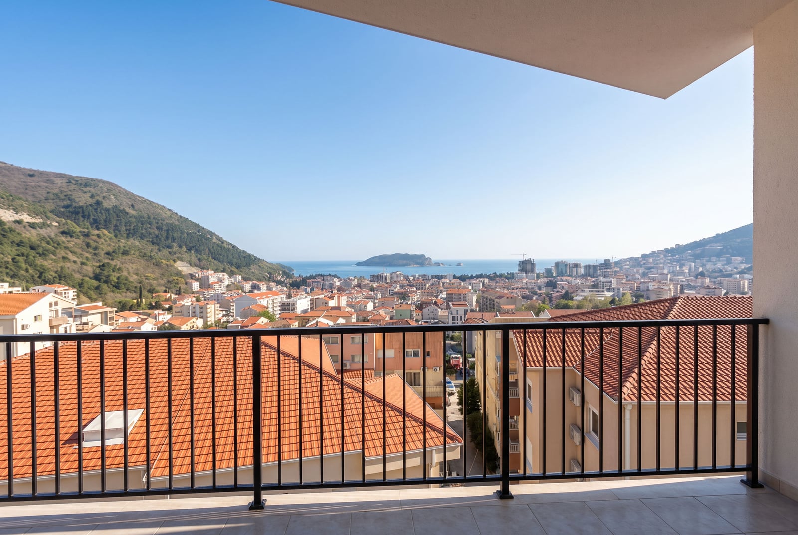 View from a balcony overlooking city rooftops, hills, and the sea under a clear blue sky.