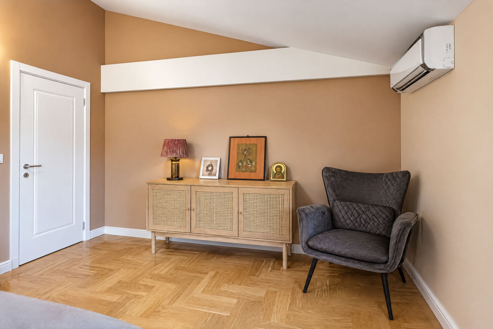 Bedroom corner with a gray armchair, wooden cabinet, and parquet floor. Part of a bed and AC unit visible.