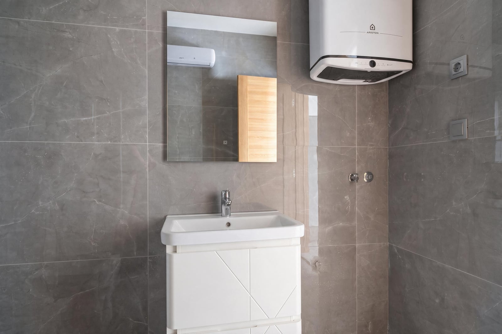 Modern bathroom with gray tiles, white vanity, mirror, and water heater in an apartment in the city of Bar, Montenegro.